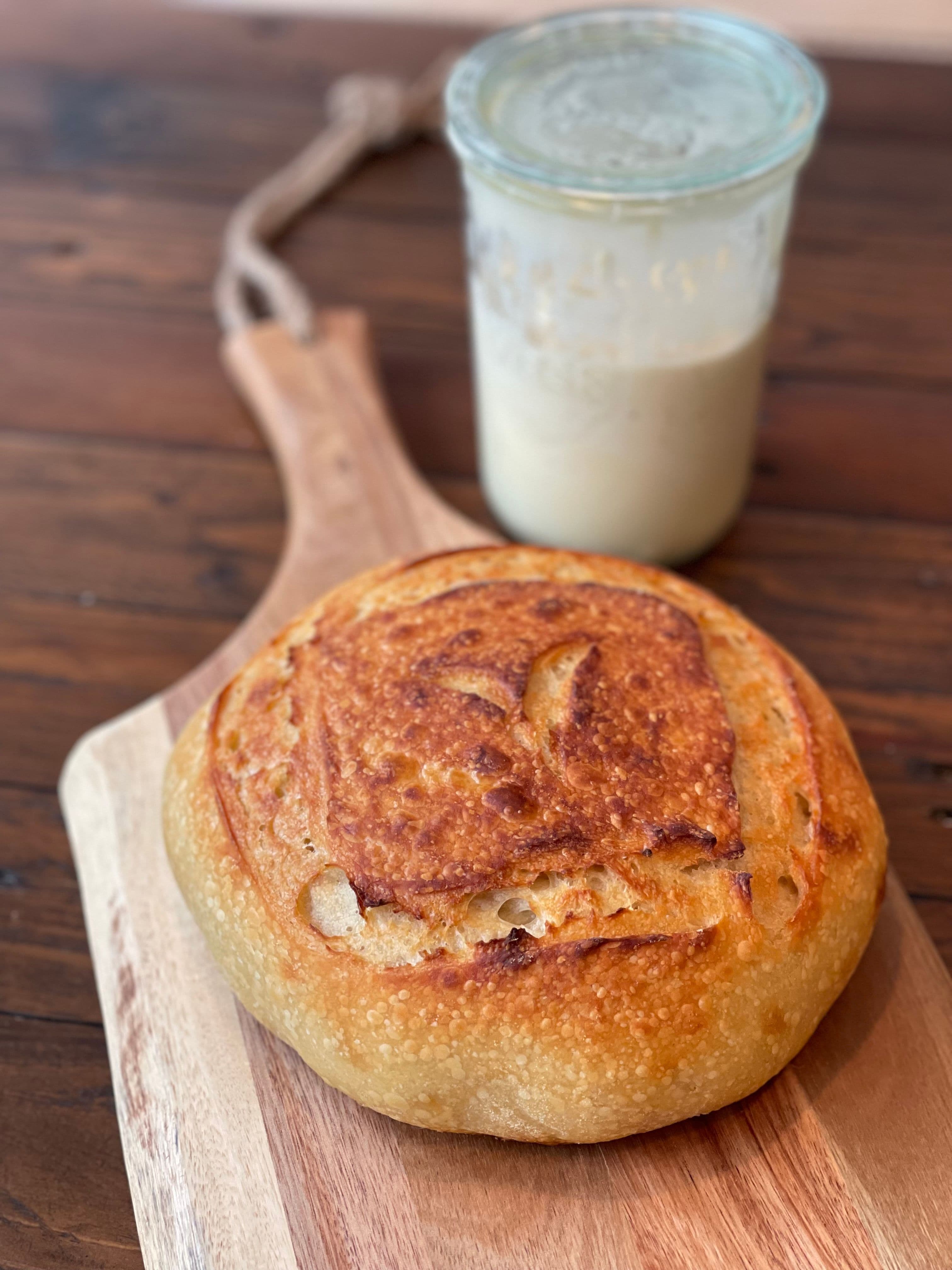 A baked loaf on a wooden board with a glass of starter behind it